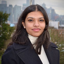 A young woman in a dark jacket and white turtleneck looks proudly at the camera.