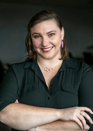 PhD Candidate Emily Sample stands with her arms crossed and smiles at the camera. She is wearing a black collared shirt and a golden necklace with three interconnected circles.