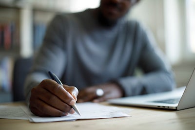 man writing with pen by a laptop