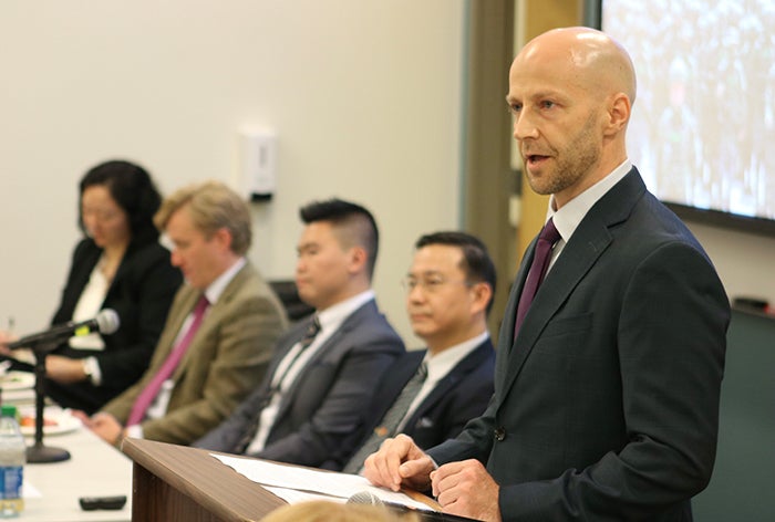 A bald man at a podium addresses an audience while four people sit in chairs to his right.