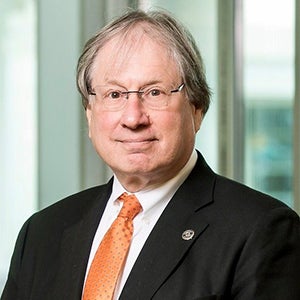 A man in eyeglasses, a black jacket, orange necktie, and thinning hair stands in front of a greenish background.