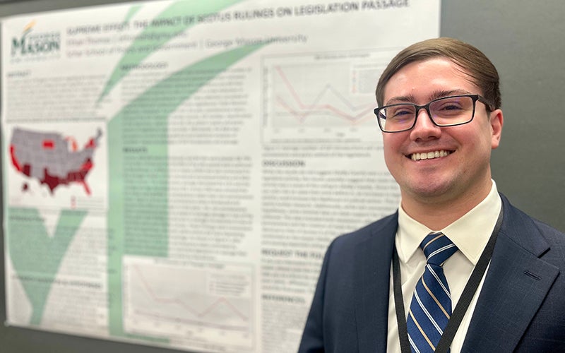 A young man wearing glasses and a navy suit jacket and tie stands in front of a research poster.