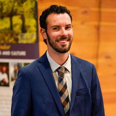 A young man with dark hair and a beard wearing a blue suit smiles at the camera.