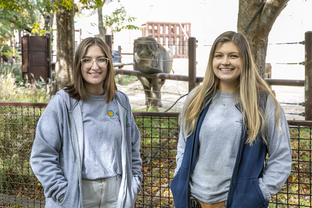 Smithsonian-Mason School of Conservation student Eva Noroski and Mason alumna and Elephant Trails keeper Ashley Fortner stand side-by-side in front of an elephant enclosure at the National Zoo.   A small Asian elephant resting her trunk on the enclosure railing peeks in between the two women.
