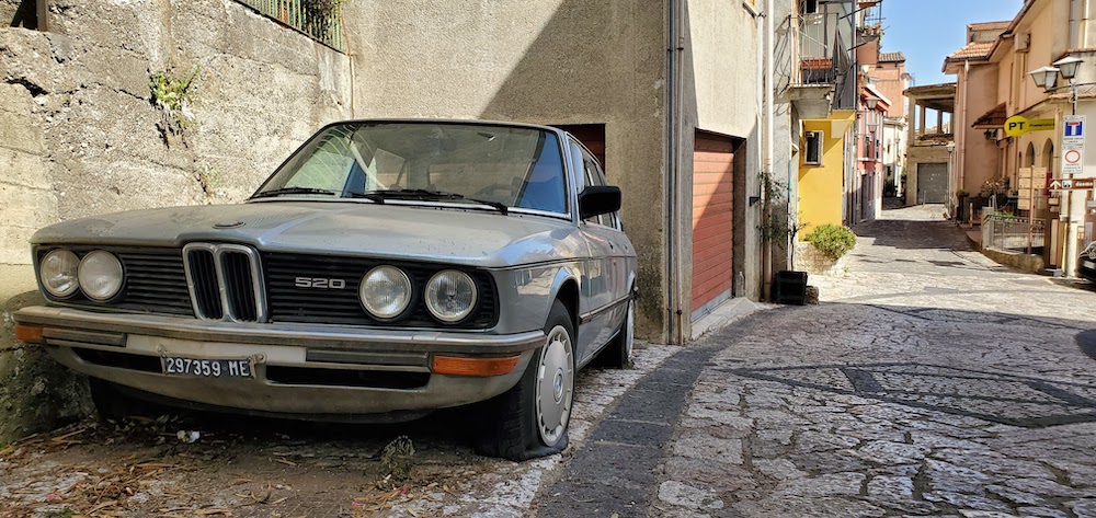 A gray BMW from the 1980s parked on the left side of a European alleyway with deflated tires.
