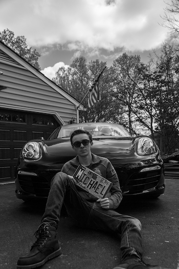 A black and white portrait of Mason student Michael Riggi sitting in front of his car, wearing sunglasses, and holding a license plate that says "Michael."