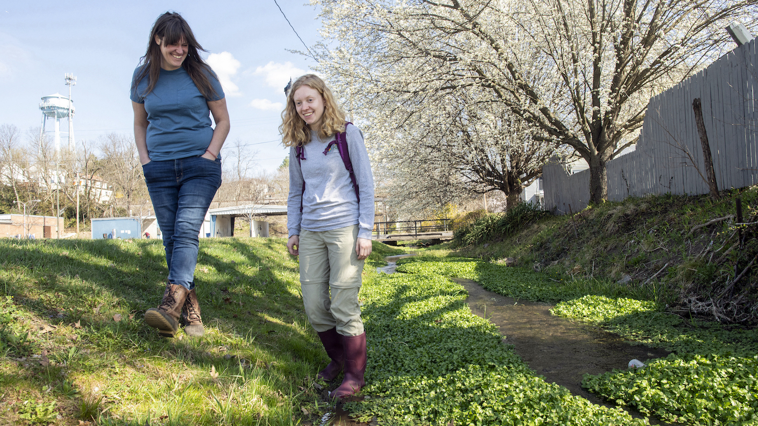 Julia Sargent and Maggie Walker walk side by side on the grass near a creek near a city street.