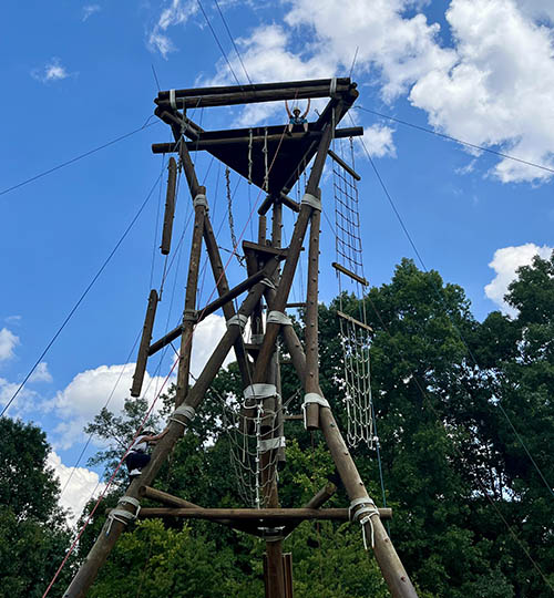 A person sits atop a tower made of logs and ropes while another in a white shirt climbs.