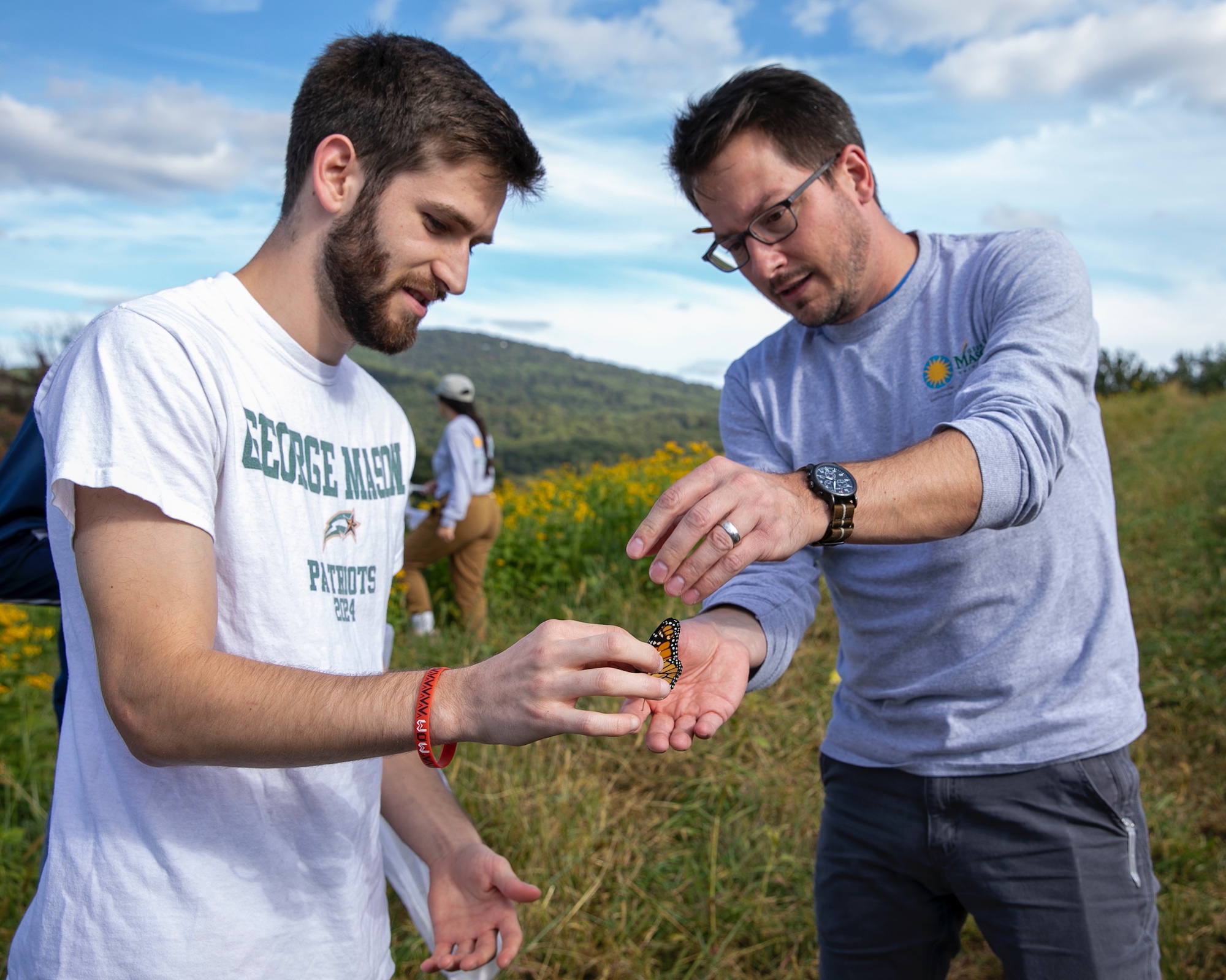 An SMSC student wearing a George Mason University T-Shirt holds a monarch butterfly while Professor Joshua Davis reaches his hand out to explain how to tag the butterfly on its hind wing.