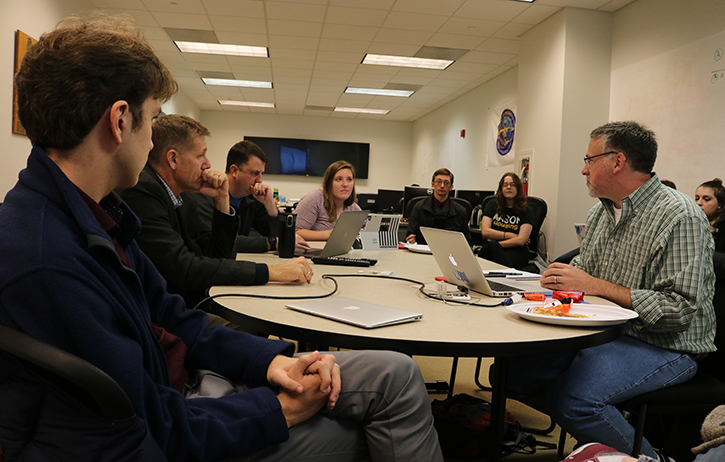 Students and faculty sit gathered around a round table with laptops.