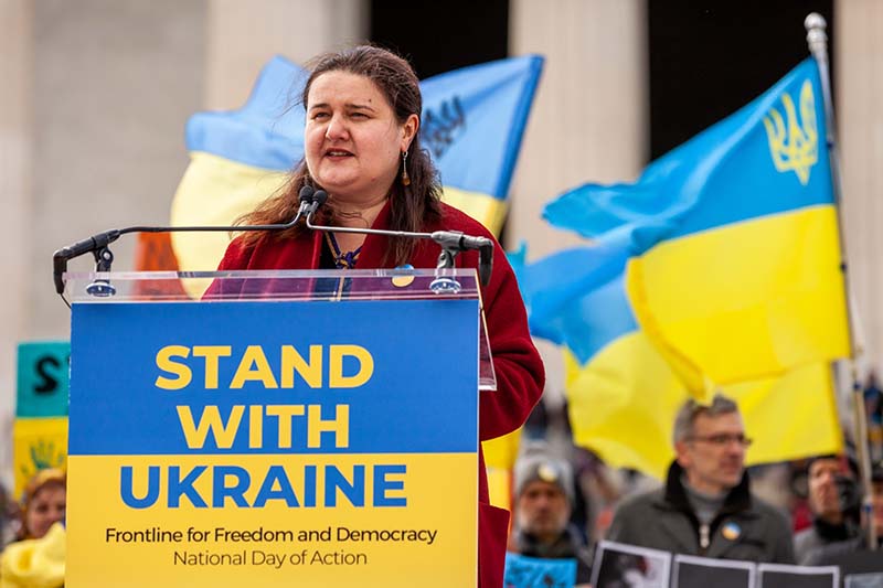 A woman surrounded by yellow and blue flags speaks into a microphone behind a podium with a sign saying Stand With Ukraine.