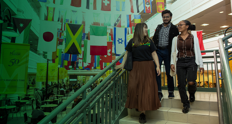 A group of three students walks down a set of stair inside the Johnson Center, which is decorated with national flags from around the world.