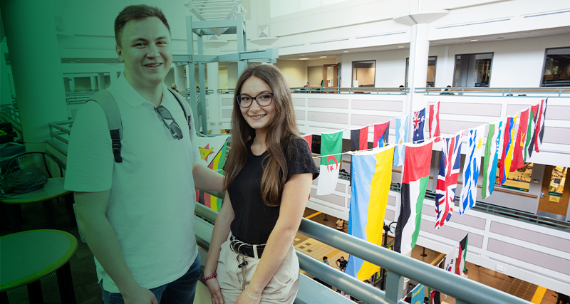 A pair of students stand on a balcony in the Johnson Center, which is decorated with international flags