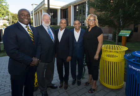 A group of people pose for a photo in front of Fenwick Library. Beside them is a yellow trashcan with a plaque, which serves as tribute to the Ahmed brothers' success.
