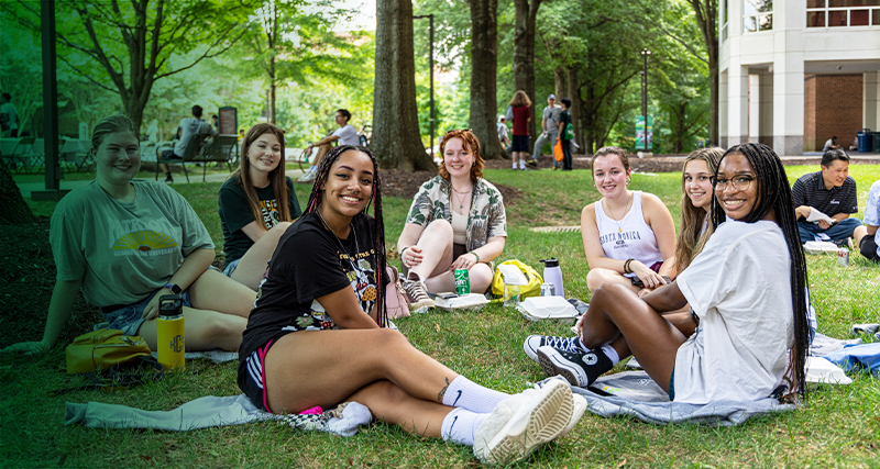 A group of students enjoy the weather while sitting together under the trees on the lawn next to Wilkins Plaza and the Johnson Center.