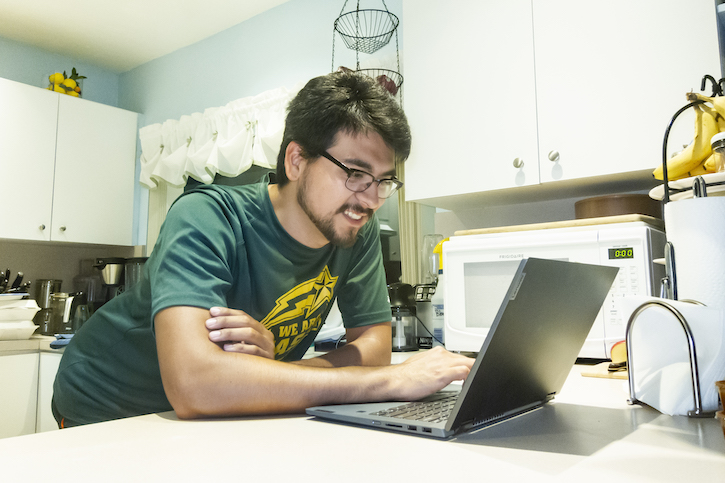 Man stands at kitchen island working on laptop. He's wearing a George Mason University t-shirt and looks engaged in his work and relaxed.