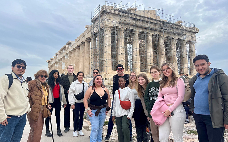 A group of people in jackets, except for one woman in a sleeveless top, stand in front of the Parthenon in Athens. It is covered in scaffolding.