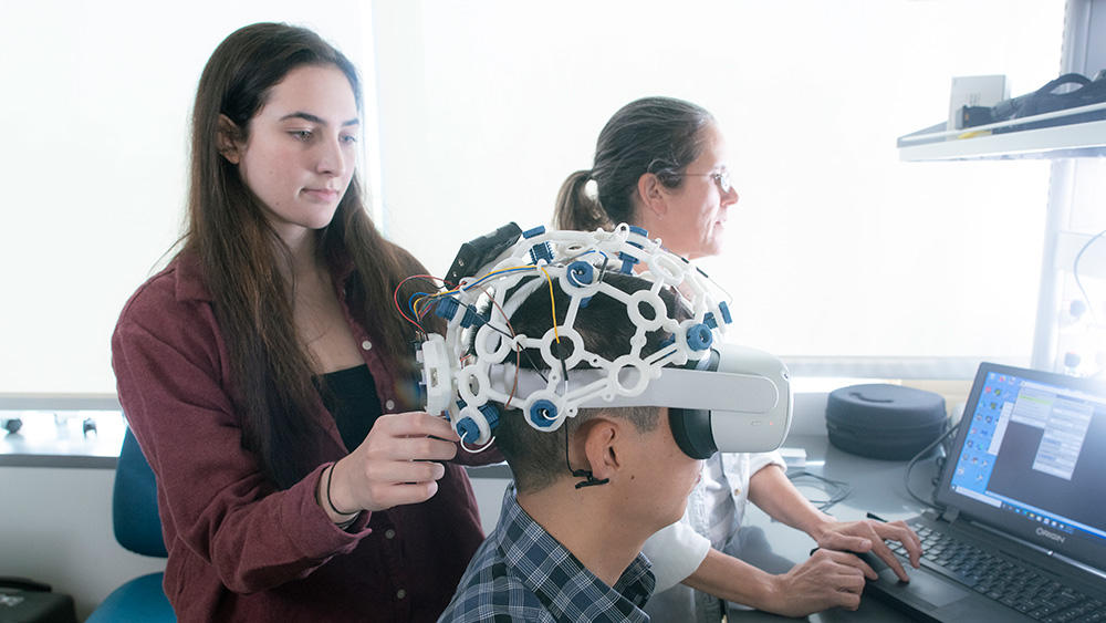 A researcher places a neural mapping device on a person already wearing a VR headset. In the background, another researcher is looking at a computer.