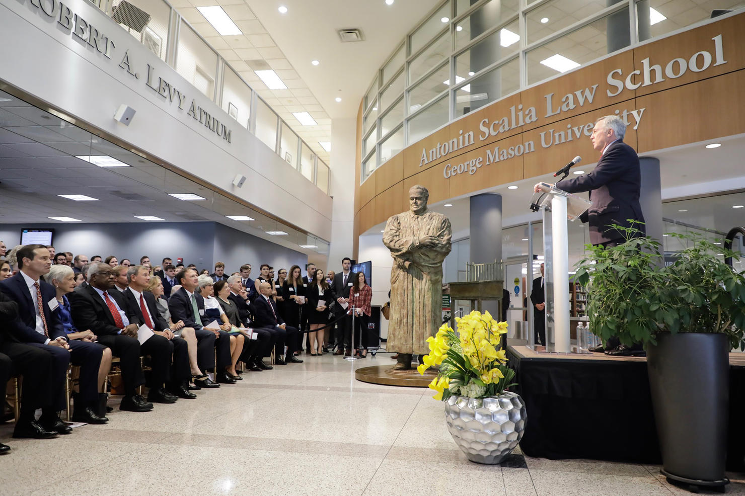 The Scalia statue is unveiled in the center atrium of the law school. To the right, the dean is seen addressing the audience at a podium, and the audience, including five Supreme Court Justices are sitting in rows to the left.