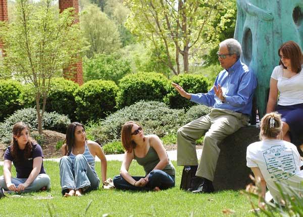 Roger Wilkins rests on the plinth of &quot;Communitas&quot; environmental sculpture on the Fairfax campus of George Mason University. He is speaking to a group of students. It is a beautiful sunny spring day, and students listen attentively as they are seated on the ground. Wilkins is wearing khaki trousers, a blue oxford shirt, and sunglasses.