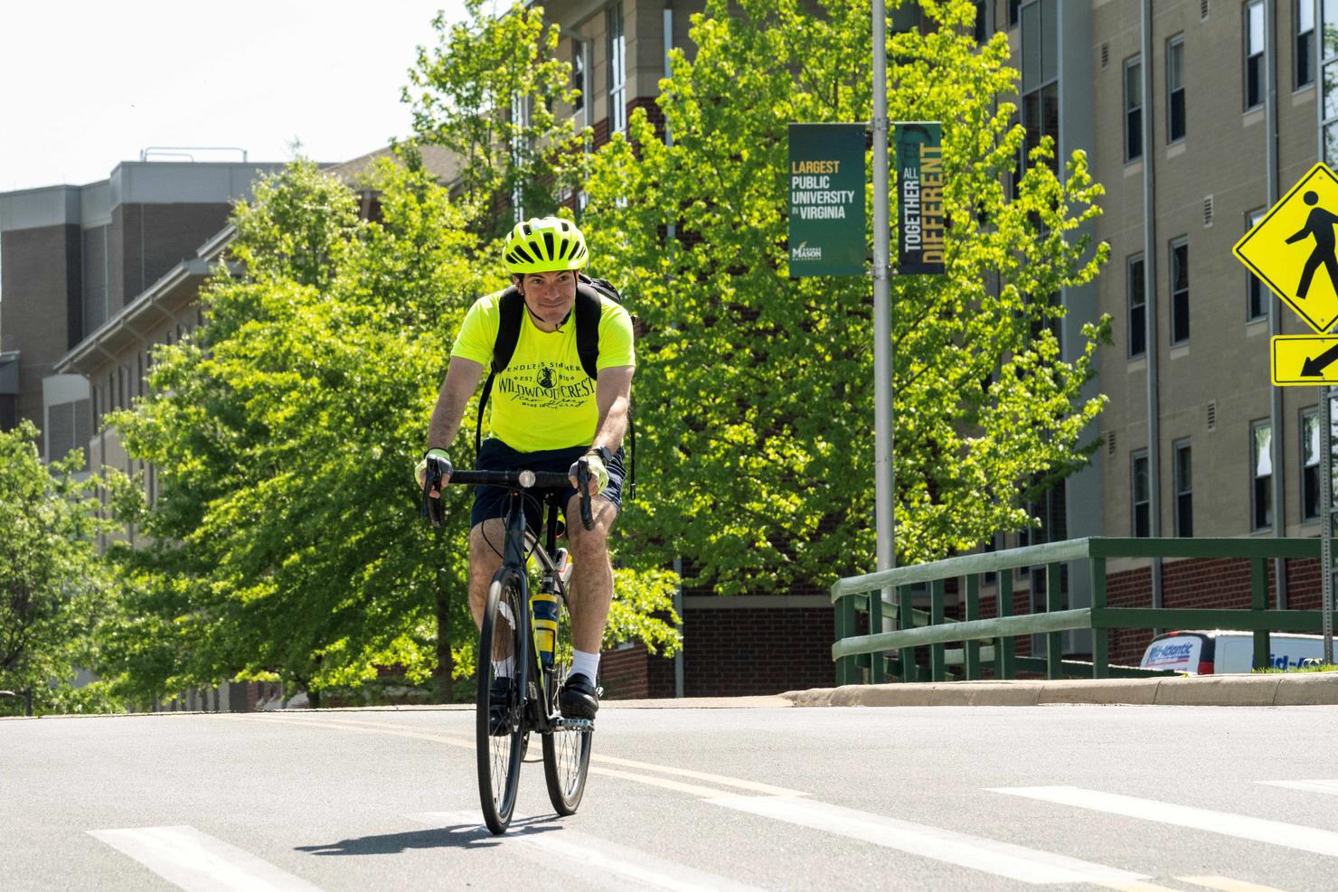 Kenneth Strazzeri rides his bike on the Fairfax Campus of George Mason University. The background includes a brick building and a light pole banner that says Largest Public University in Virginia, and All Together Different