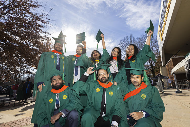 A group of masters students pose for the camera, their caps held high in the air.