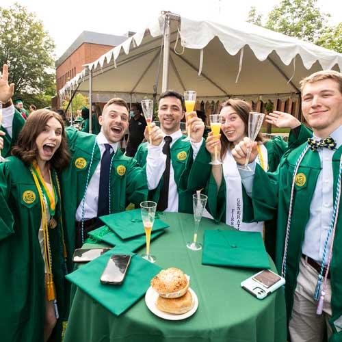Graduates enjoy bagels and bubbles, a an alumni association tradition. Graduates are gathered around a table, where bagels are shown. They raise a glass of orange juice in champagne flutes in a toast to  their achievements.