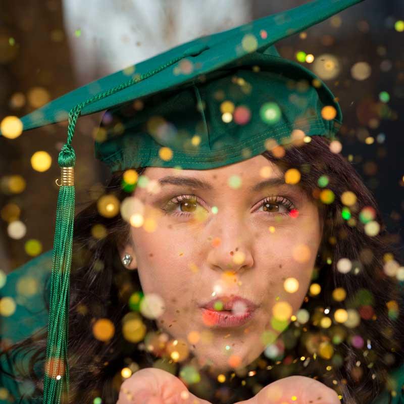 A graduate blows sparkly confetti at the camera lens