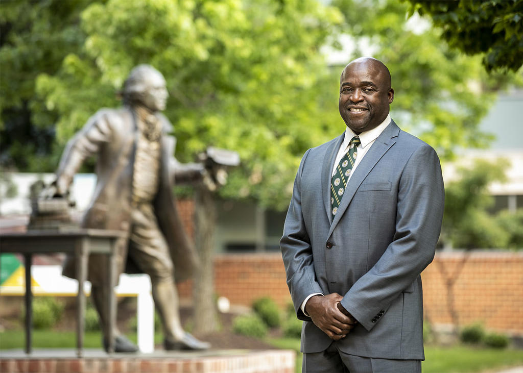 Mason President Gregory Washington stands outside. He is dressed in a gray suit, Mason logo tie. The George Mason statue is to his left.