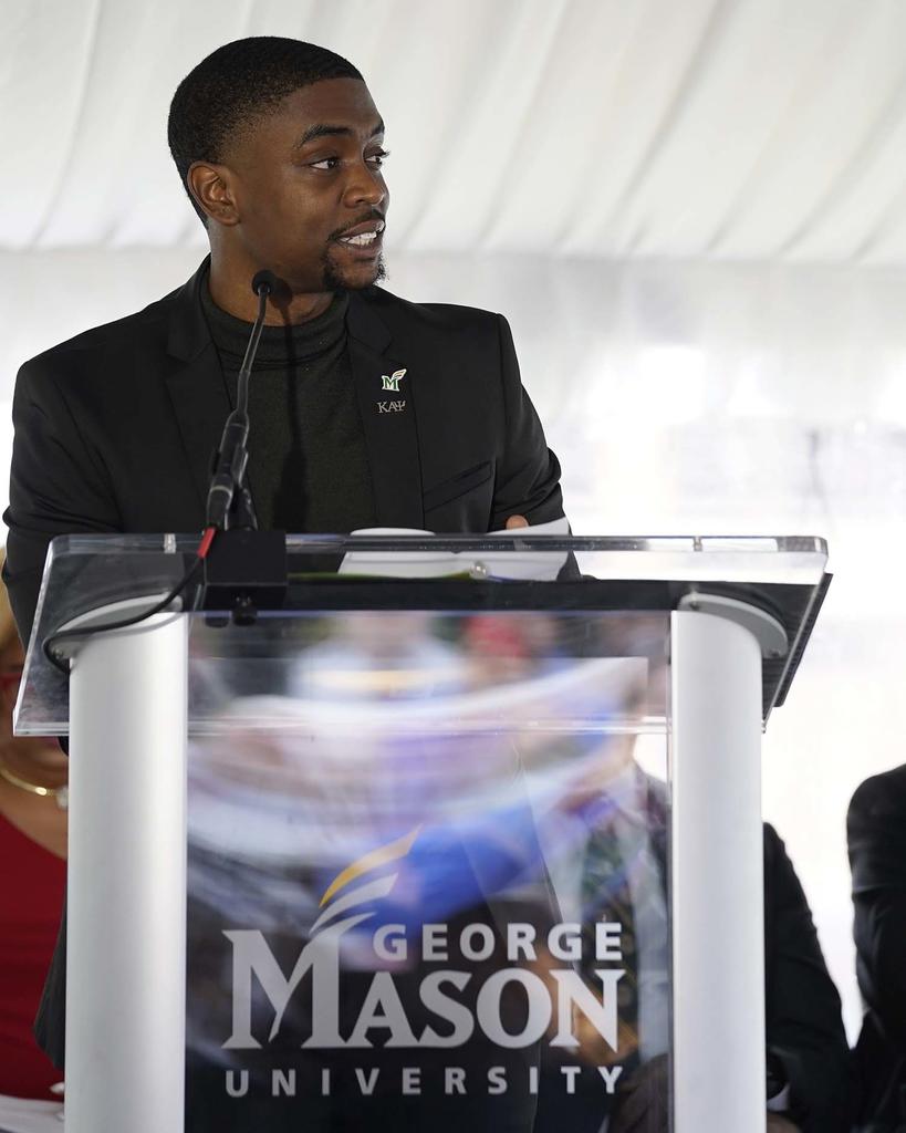 Young Black man wearing a black suit stands at a clear podium with the words George Mason University engraved on the front. 