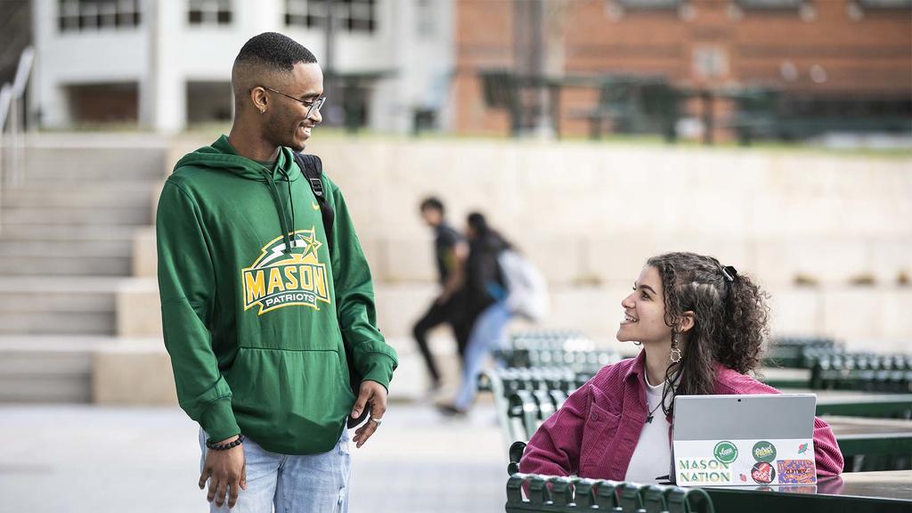 two students talking outside. A female student wearing a pink jacket is seated with a laptop on the table. She is speaking with a male student, standing, wearing a green and yellow Mason sweatwhirt. 
