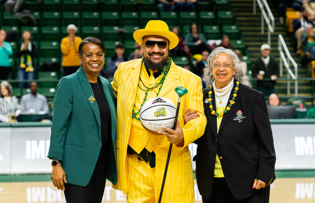 "Doc Nix" Nickens dressed in a yellow suit, green mardi gras beads around his neck, wearing a yellow hat, holding a basketball. Athletic director Nena Rogers to his left, his mother Linda West to his right. They stand on the court as Doc Nix is honored by Mason Athletics