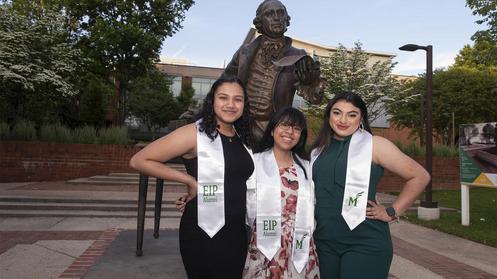 Three female students posing outside in front of the statue of George Mason, each wearing a white sash/scarf with the letters EIP short for Early Identification Program.