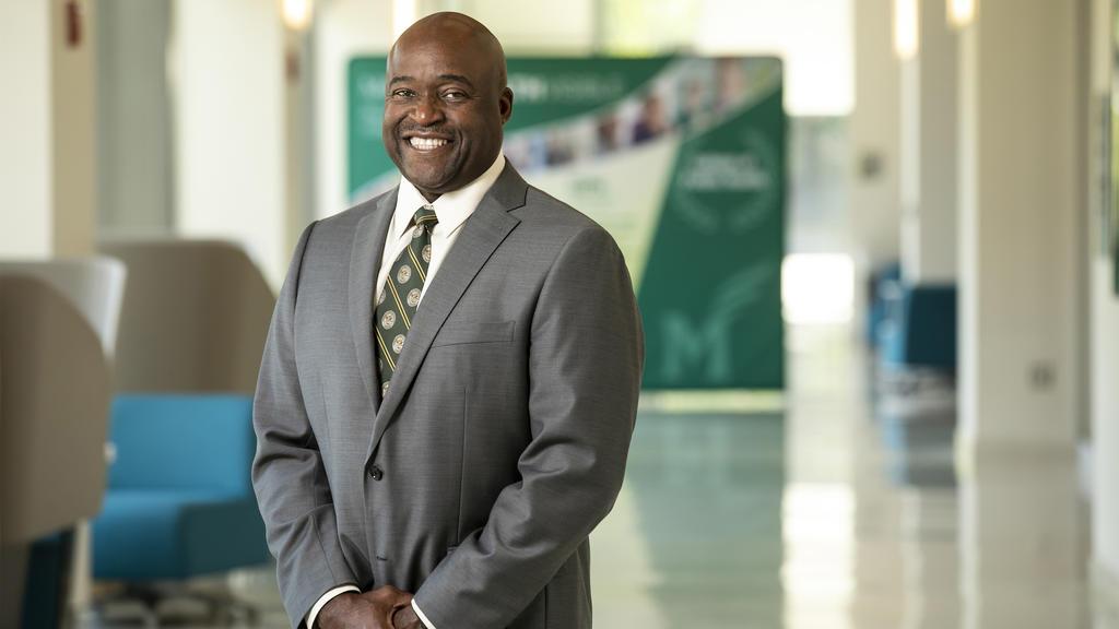 Mason president, wearing a gray suit and green and gold tie, smiles for the camera in a light filled building atrium.