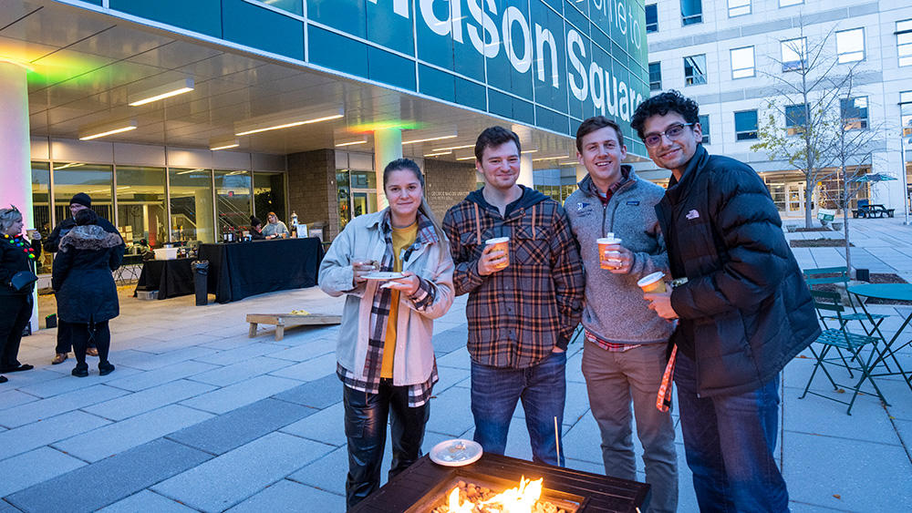 Students stand on plaza at mason square enjoying hot beverages by a fire pit.