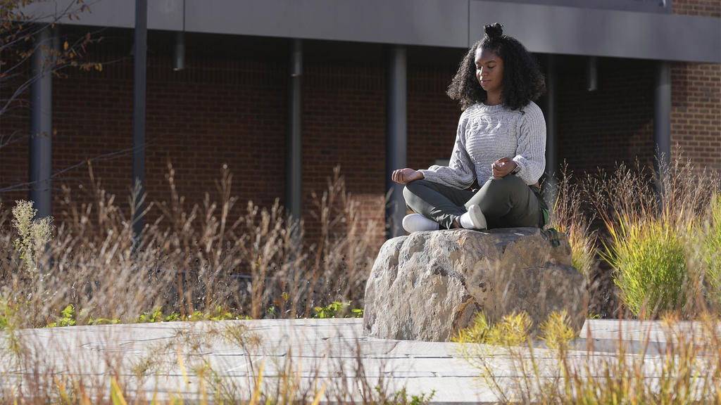 a young Black woman is meditating on a large rock or boulder on campus