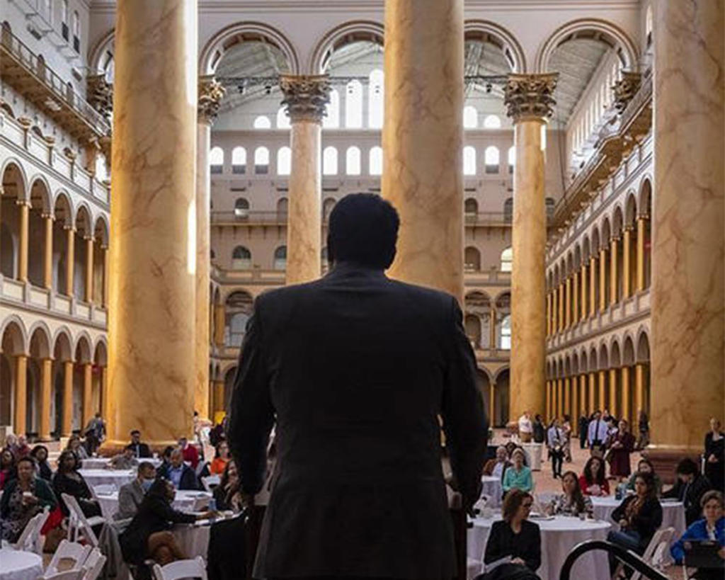 The back of a man in silhouette speaking to an audience at the National Building Museum, in the grand hall.