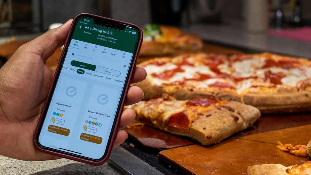 A hand holds a phone in front of pepperoni pizza. On the phone is the Everyday app. It is open to the Ike's dining hall menu, specifically showing that there is pepperoni pizza on the menu, how long it will be on the menu, and dietary information.