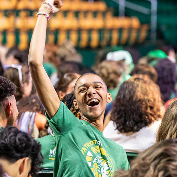 A student in Mason attire cheers, his fist thrown up into the air, amidst a crowd of other students.