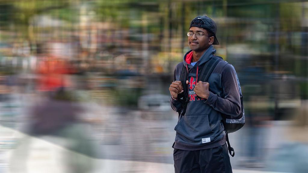 Freshman finance major J. J. Powell is in focus in the photo, the background is blurry. JJ wears a baseball cap, a gray and red sweatshirt. He has a backpack on his shoulders.