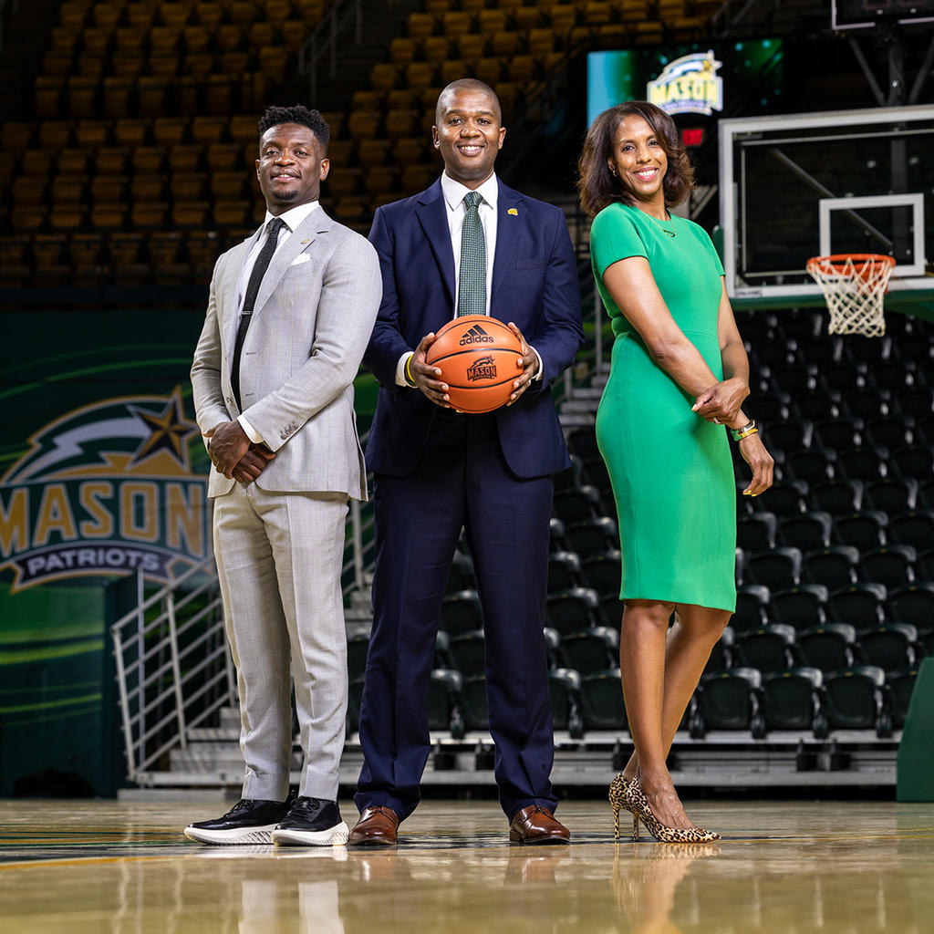 Three people stand in business attire on the Mason basketball court. The middle person holds a basketball.