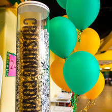 Large plexiglass column contains used vials of the COVID-19 vaccine administered at Mason. The column is decorated with the words "Crushing Covid" and there are green and gold balloons nearby.
