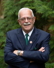 U.S. Representative Gerry Connolly in a blue suit and green tie standing with his arms crossed in front of evergreen trees.