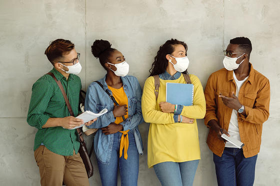 a group of four multiethnic students standing against a wall all wearing protective masks