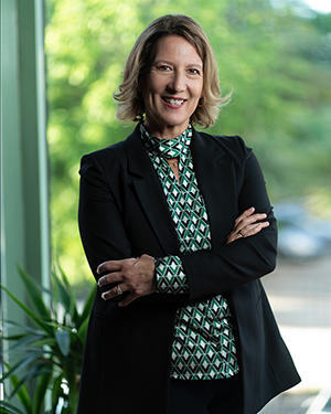 Melissa Perry wears a green and blue print blouse and a dark jacket while standing in front of a window with trees outside
