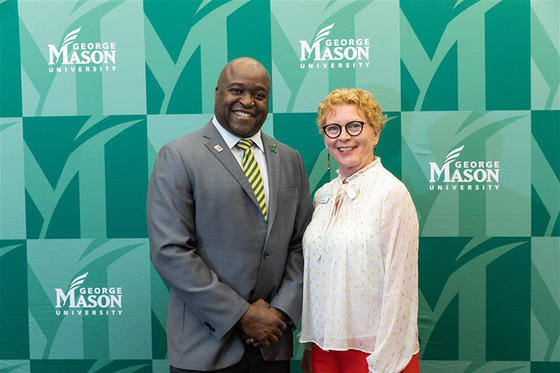 Gregory Washington in a gray suit and green and gold striped tie stands next to Janet Gullickson in a white blouse and red pants