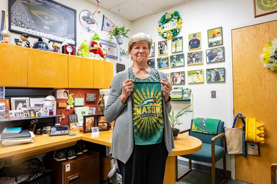 Jeanne Medford stands in her Mason Athletics office, holding a Mason flag. The office is decorated in Mason and New York Yankees memorabilia.