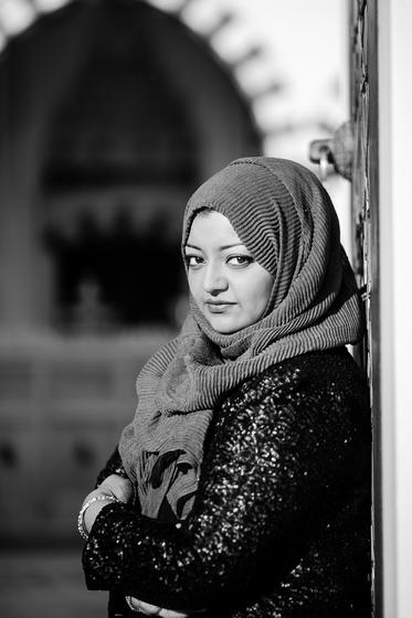 Portrait of Rabia Chaudry. Woman wearing head scarf leans against the iron gate of an old building, gazing at the camera thoughtfully.