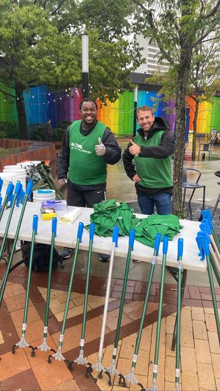 Micah Hodges, at left, stands at a table outdoors with a fellow participant in NOVA Cleanups. They have materials and supplies at the table to help volunteers clean up trash.