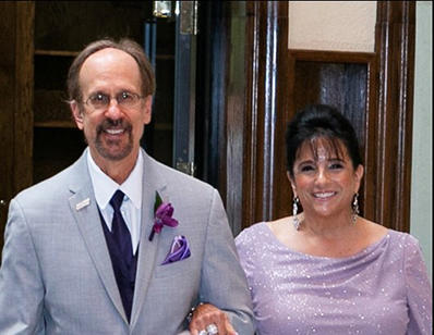 Greg Baroni, with glasses and a goatee,  is wearing a grey suit with a purple tie, boutonierre and pocket square. Camille Baroni wears a lavender gown.